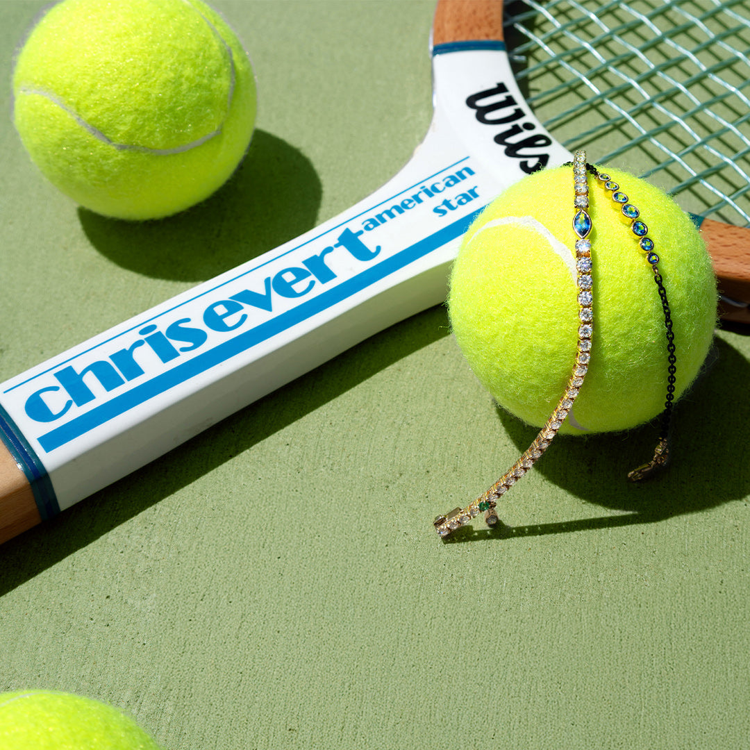 A tennis racket with a white grip printed with 'chrisvert' and 'Williams' lies on a green tennis court surface, alongside several bright yellow tennis balls. A delicate rhinestone tennis bracelet is draped over one of the balls.
