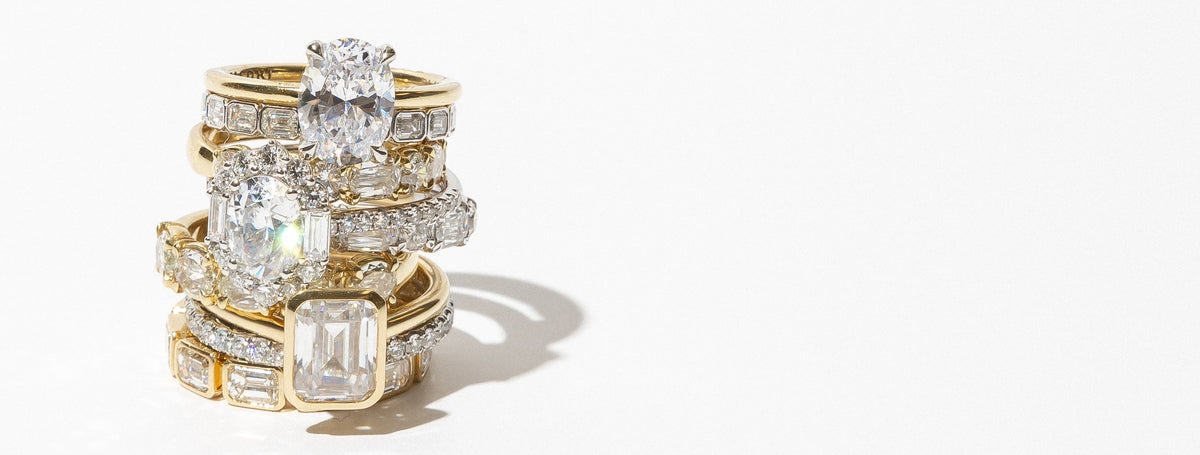 A stack of gold rings adorned with multiple large clear gemstones, including emerald-cut and round diamonds, photographed on a clean white background with a soft shadow.